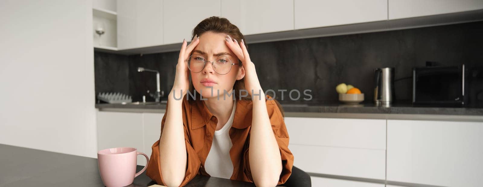Portrait of working woman at home, student has headache, touches head and has painful migraine, sits in kitchen in glasses.