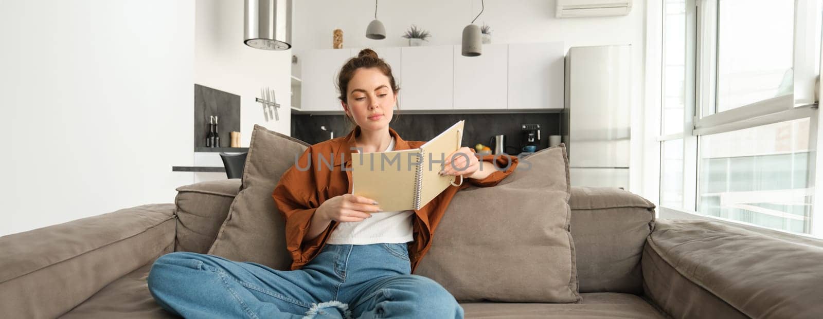 Smiling young brunette woman, sits on sofa in living room, holds notebook, reads her notes, studies for exam, student does her homework at home.