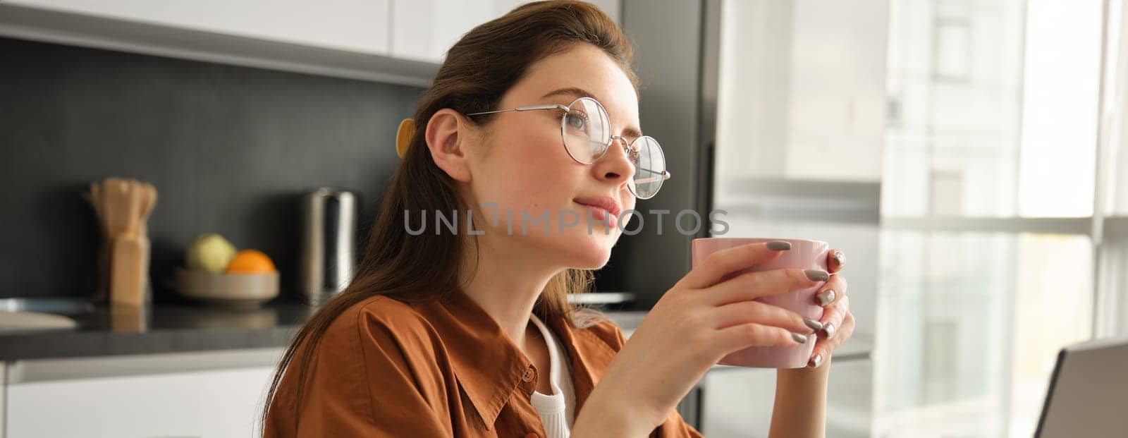 Portrait of young woman enjoys quite day at home with hot cup of tea. Female model sits in kitchen with mug of aromatic coffee, wearing glasses, looking aside.