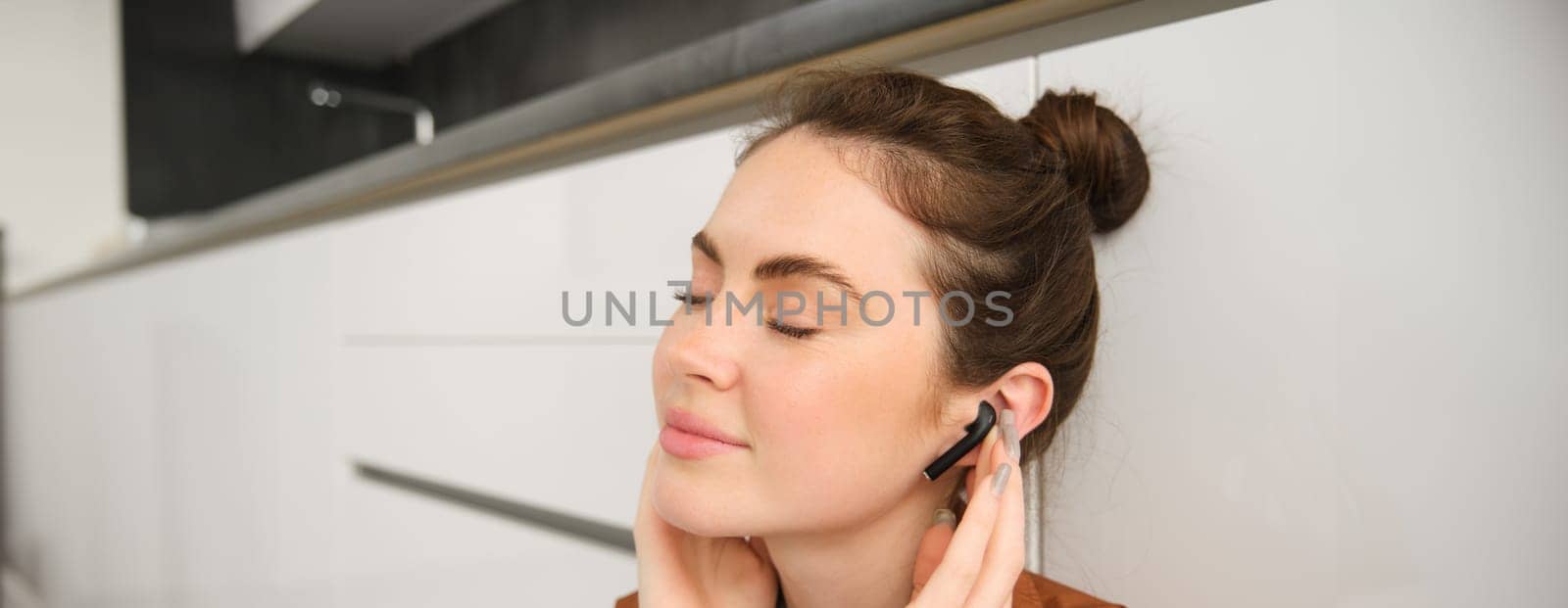 Portrait of young smiling woman listens to music in her black wireless earphones, using headphones to enjoy sound quality of song, sits on kitchen floor.