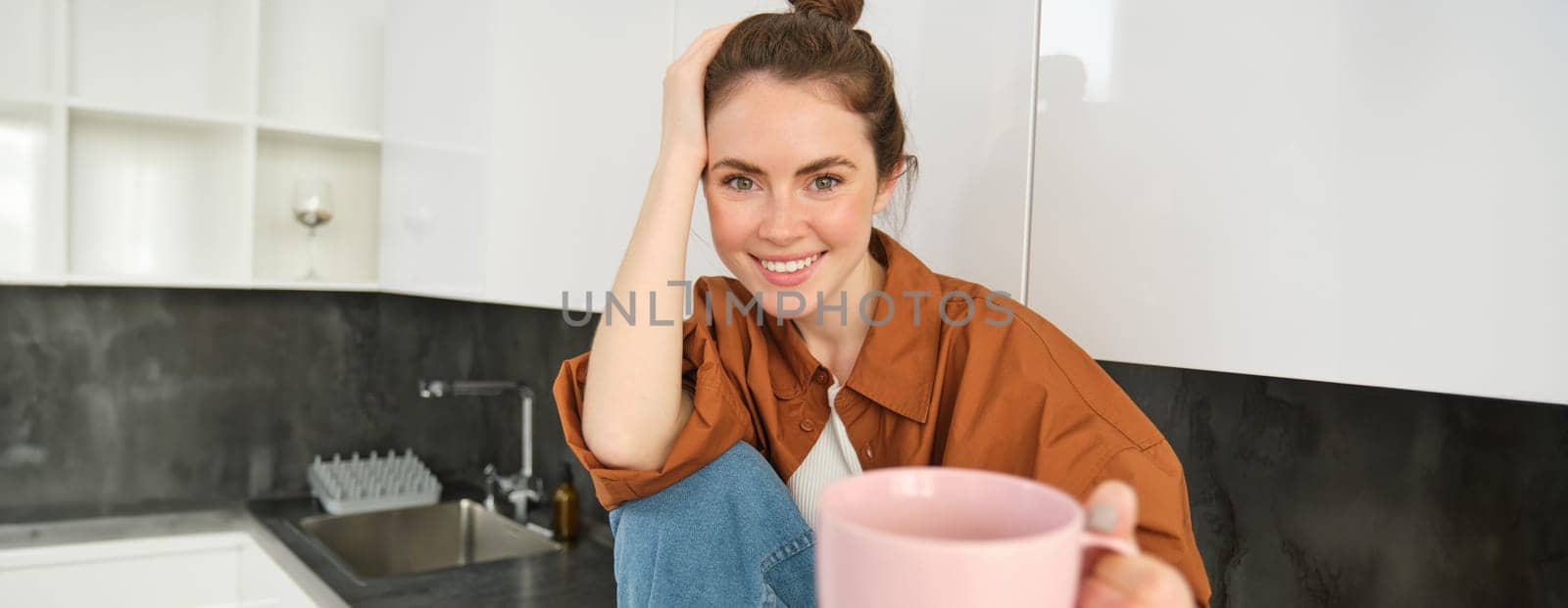 Beautiful young brunette woman sits in kitchen, drinks coffee, offers you cup and smiles, looks happy.
