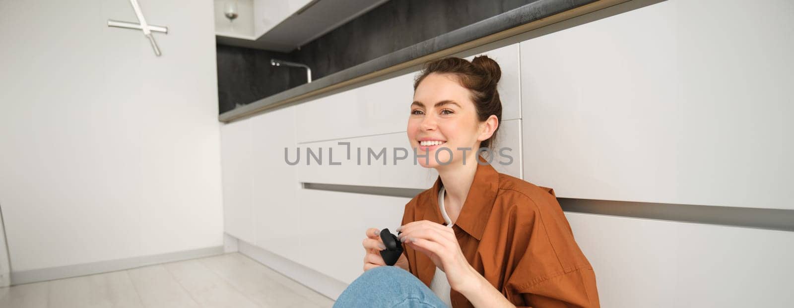 Portrait of young smiling woman listens to music in her black wireless earphones, using headphones to enjoy sound quality of song, sits on kitchen floor by Benzoix