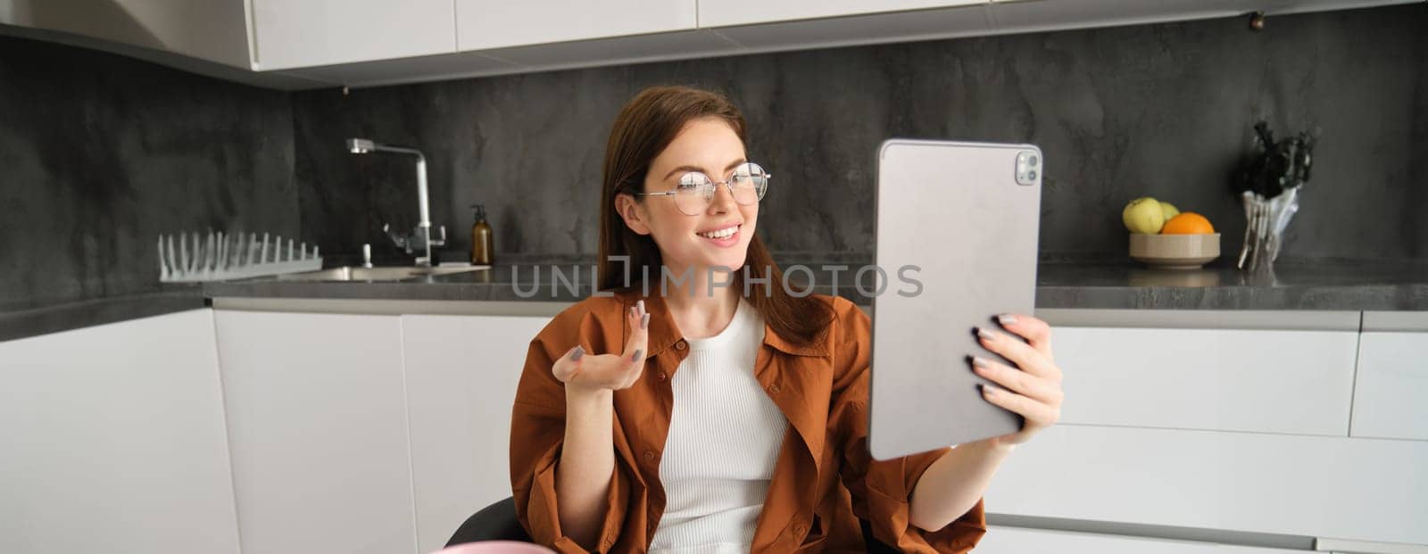 Portrait of young woman joins online course on digital tablet, having conversation, chatting on remote, sitting in kitchen.