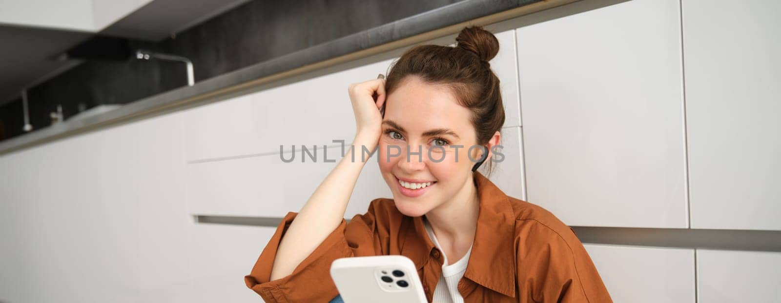 Portrait of beautiful smiling woman sitting on kitchen floor, scrolling social media on mobile phone, listening music in wireless headphones.