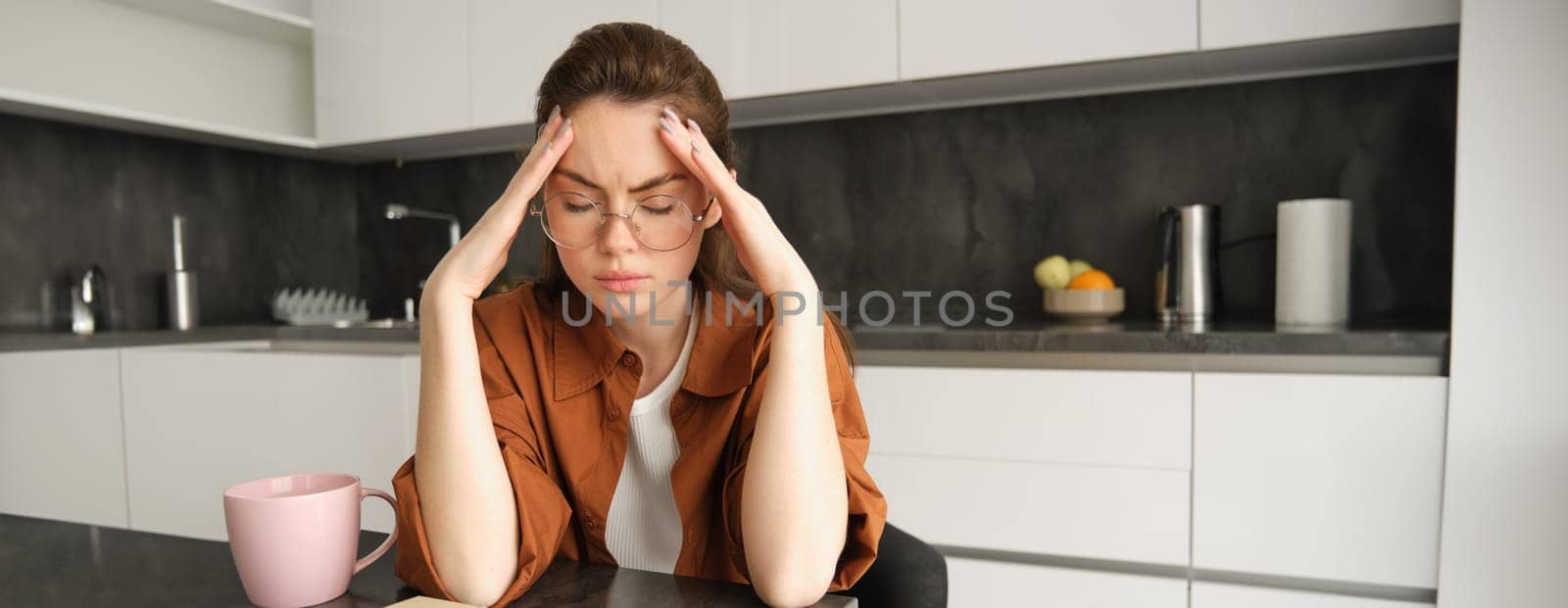 Portrait of tired young working woman in glasses, holds hands on head, suffers painful migraine, has pain in temples, sitting with headache in kitchen by Benzoix