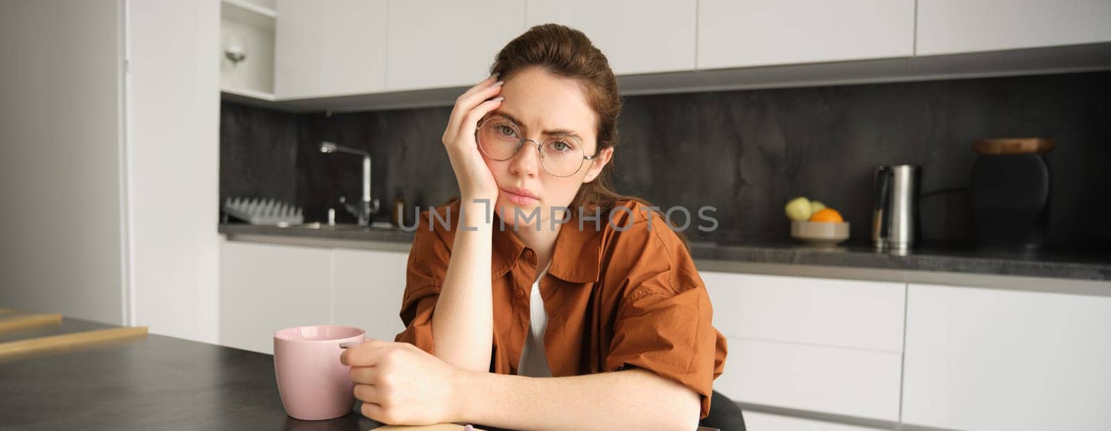 Portrait of young serious woman, working from home in glasses, sitting with folder and documents in kitchen, looking tired by Benzoix