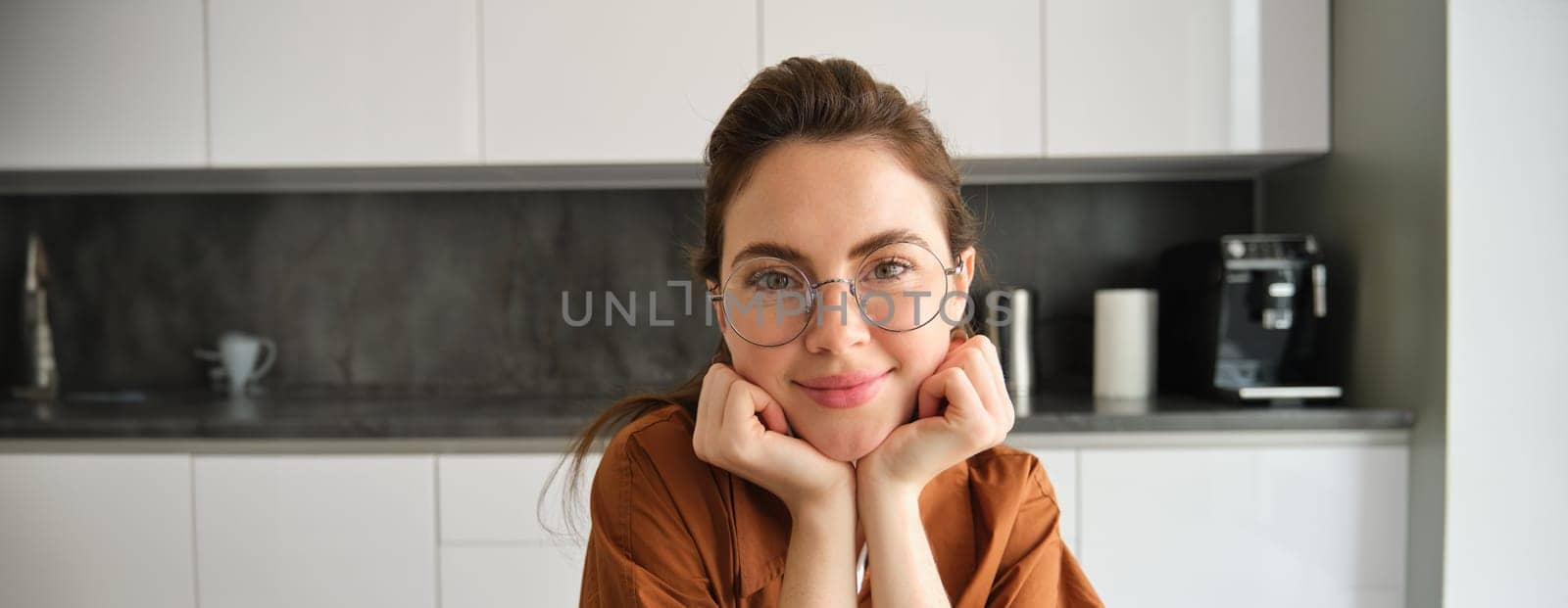 Portrait of cute, beautiful young woman in glasses, sitting at home in kitchen, lean head on hands, smiling and gazing dreamy.