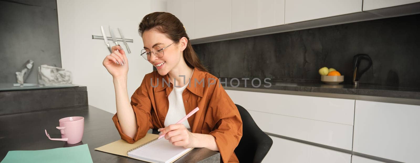 Portrait of young woman in glasses studying at home, writing down something in notebook, making notes, student doing homework in kitchen by Benzoix