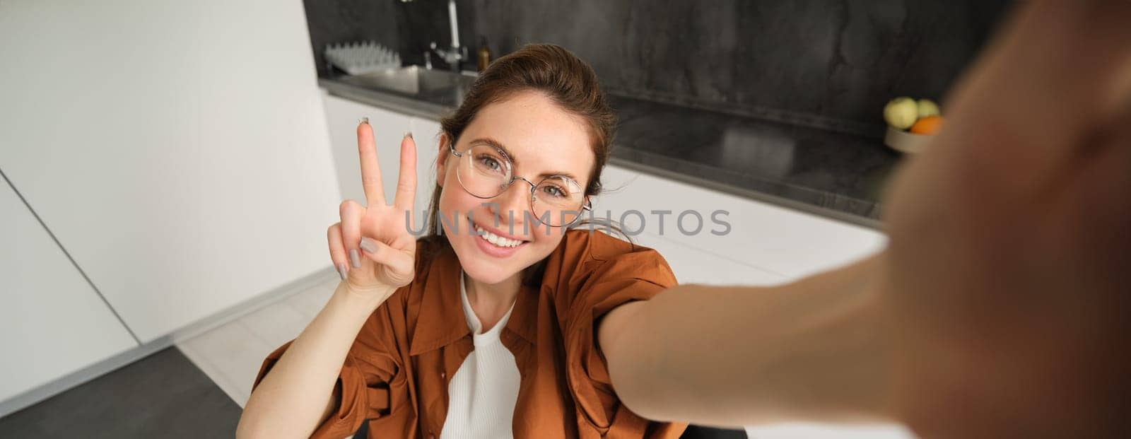 Portrait of stylish young woman at home, taking selfie on smartphone, posing in kitchen and smiling at mobile camera by Benzoix