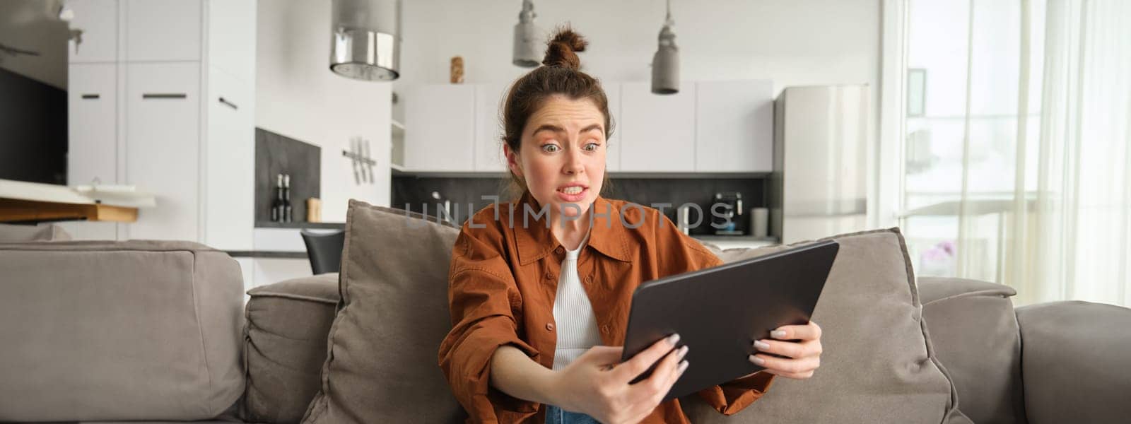 Portrait of anxious, stressed young woman looking scared at digital tablet, staring at screen, sitting on couch with gadget in living room.