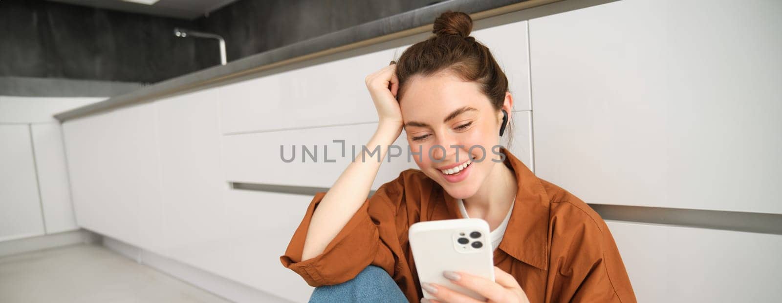 Portrait of young modern woman in wireless headphones, sitting on kitchen floor, using smartphone, making playlist with favourite songs, listens to music.