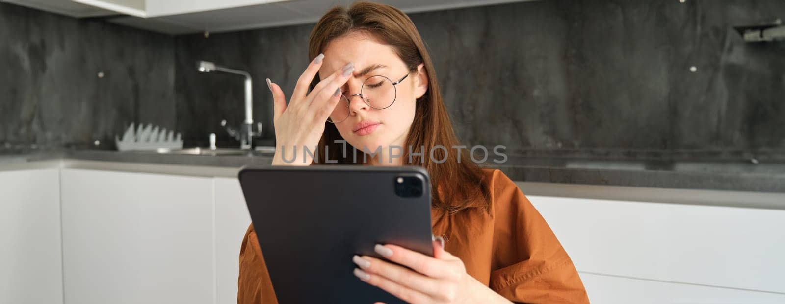 Portrait of young woman feeling unwell, touching head, has headache or migraine, reading on digital tablet in glasses.