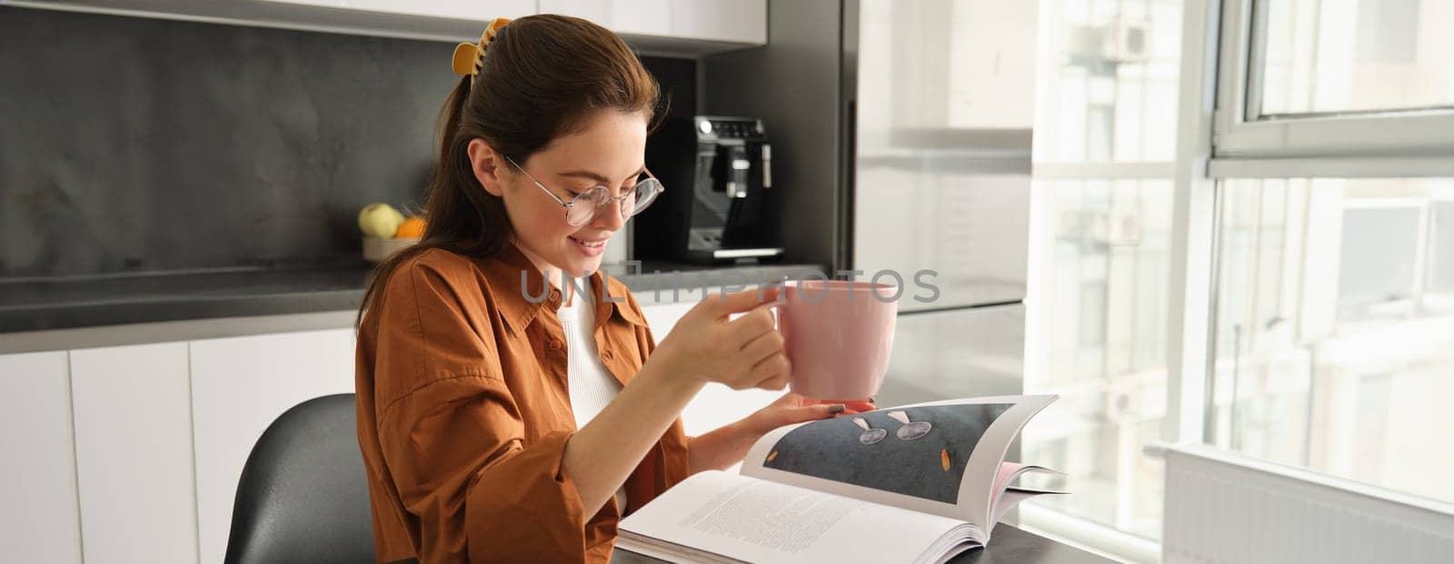 Portrait of woman reading at home, flipping pages of favourite book, relaxing in kitchen with cup of coffee, wearing glasses.
