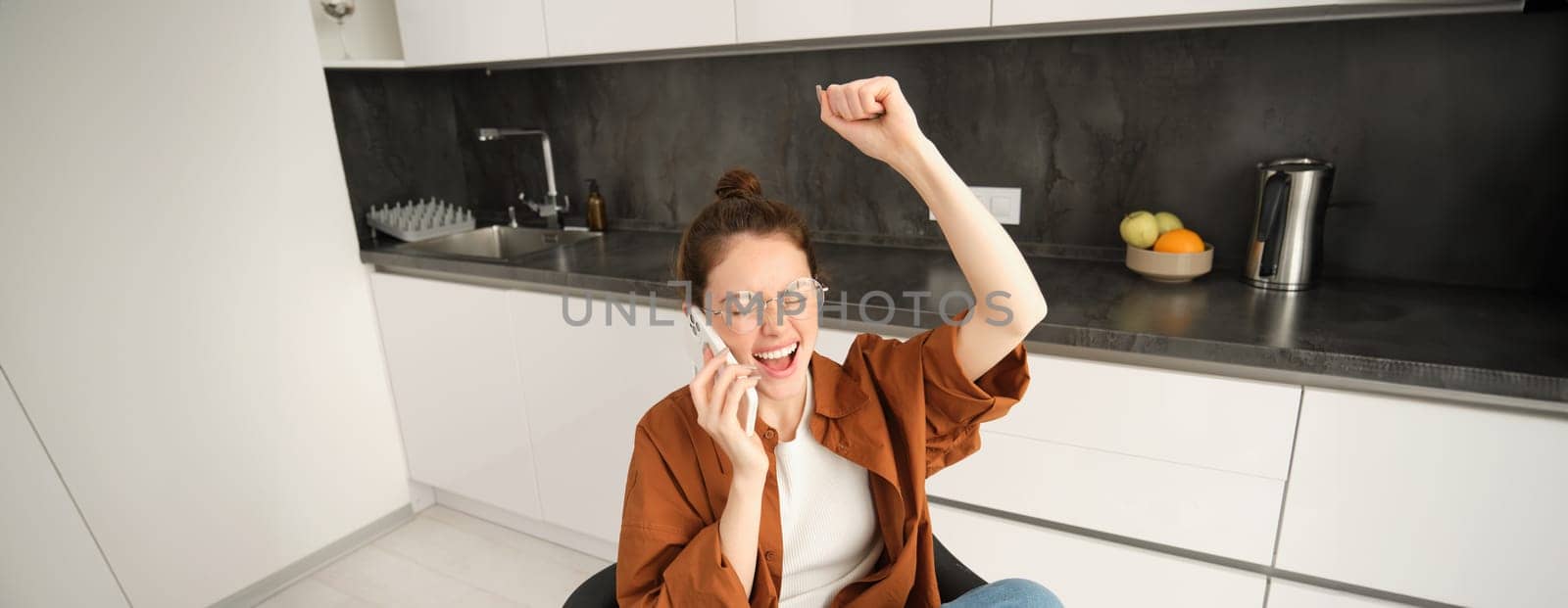 Portrait of woman receiving great news over the phone call, celebrates and jumps on chair from excitement during telephone conversation.