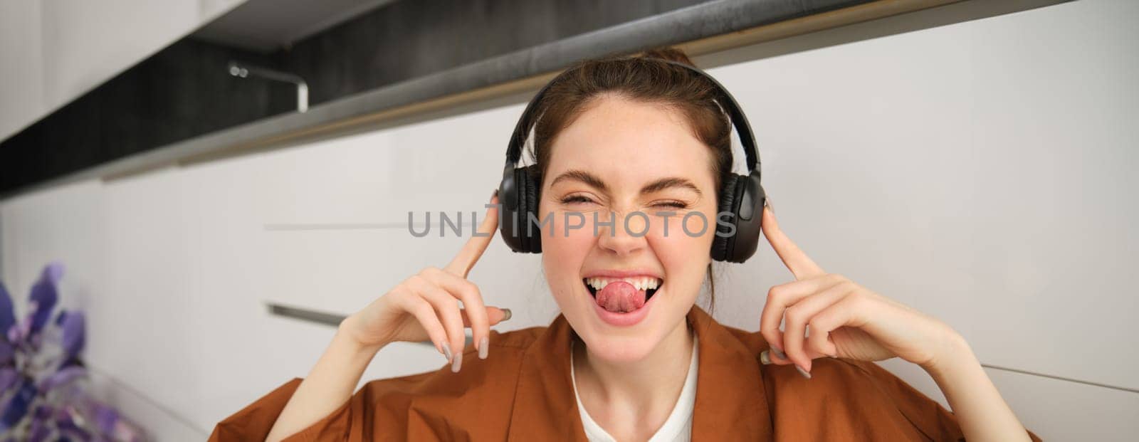 Cute young woman, makes funny faces, listens to music, wears wireless headphones, having fun at home.