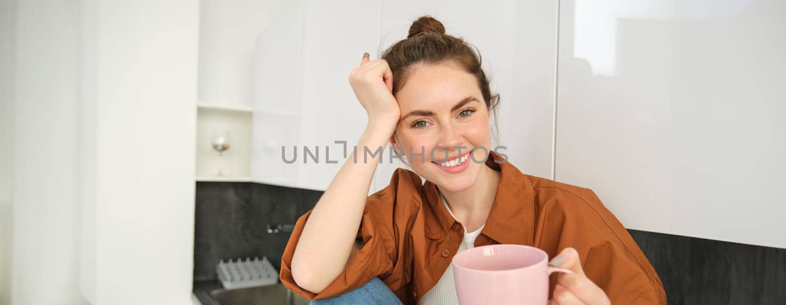 Portrait of woman with cup of coffee in hand, sits in kitchen and smiles at camera. Lifestyle and people concept