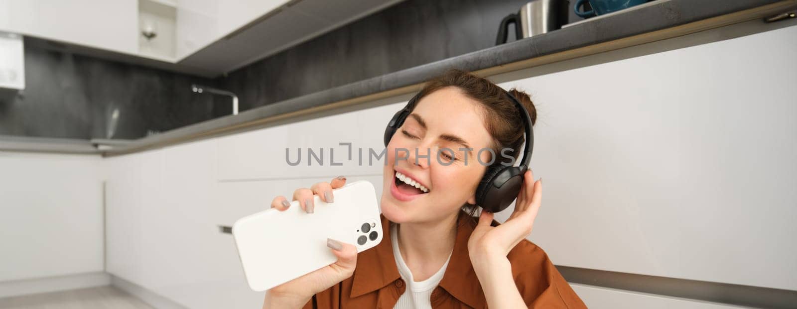 Portrait of happy, beautiful girl sings songs in headphones, listens to music, using smartphone as mic, playing karaoke app.
