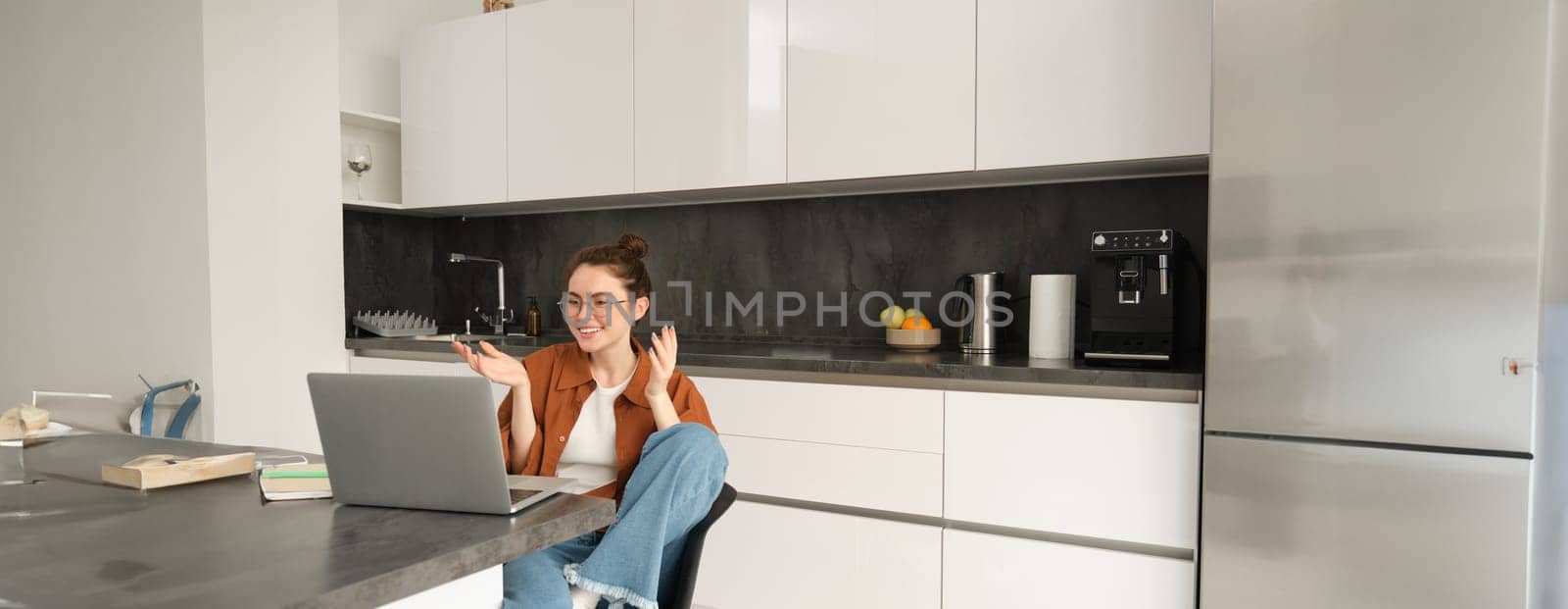 Portrait of smiling young woman, freelance tutor or teacher giving online lesson from home, connects to team meeting and working with laptop in kitchen.