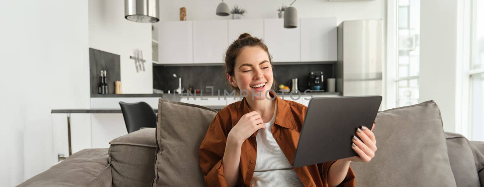 Portrait of woman laughing over funny video on digital tablet, watching movie on her gadget streaming service, sitting on sofa at home, reading e-book.