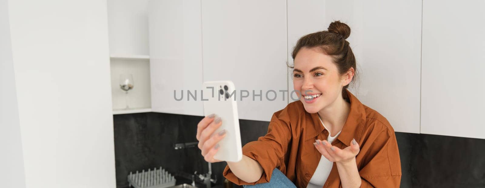 Portrait of young cheerful woman laughing and smiling during phone call, video chats with friend, sits on kitchen counter and talks to someone using smartphone app.