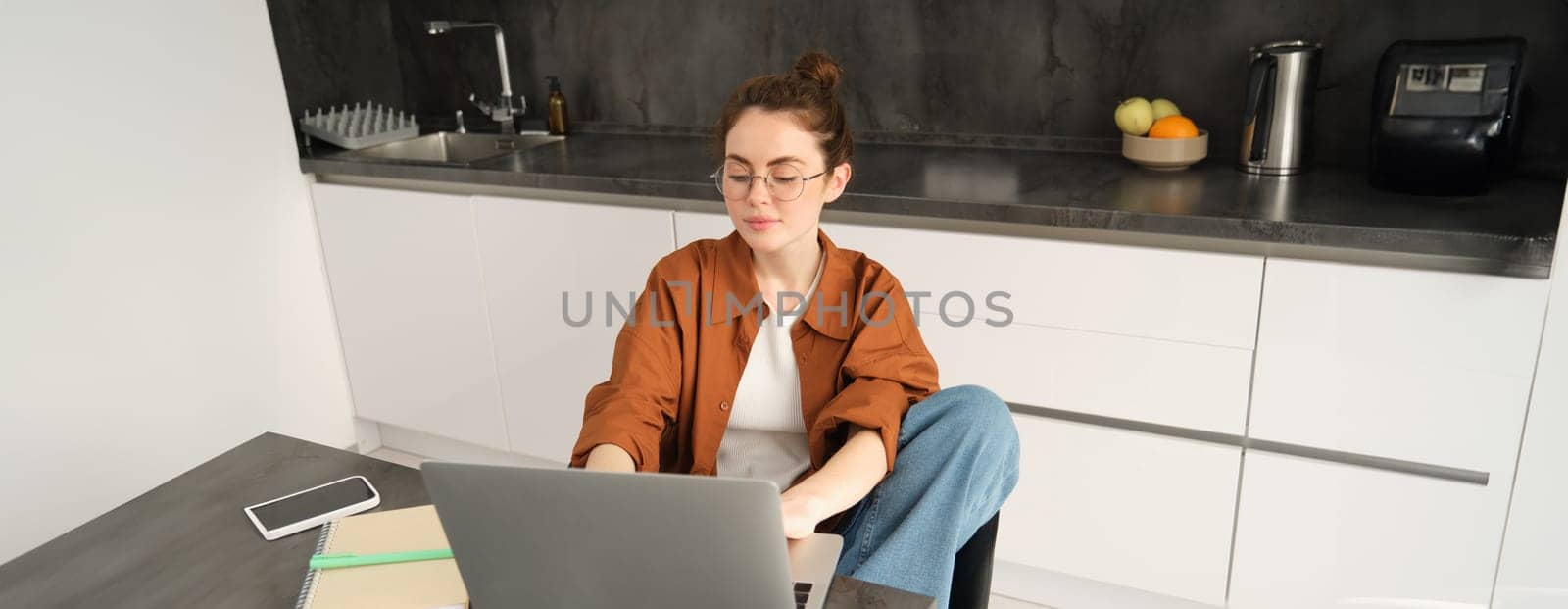 Portrait of young woman, student in kitchen, sitting with laptop, working from home, studying or doing homework, connects to online chat or course from computer.