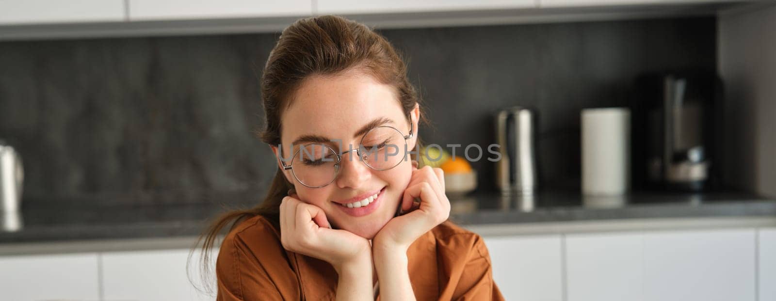 Portrait of cute, beautiful young woman in glasses, sitting at home in kitchen, lean head on hands, smiling and gazing dreamy.