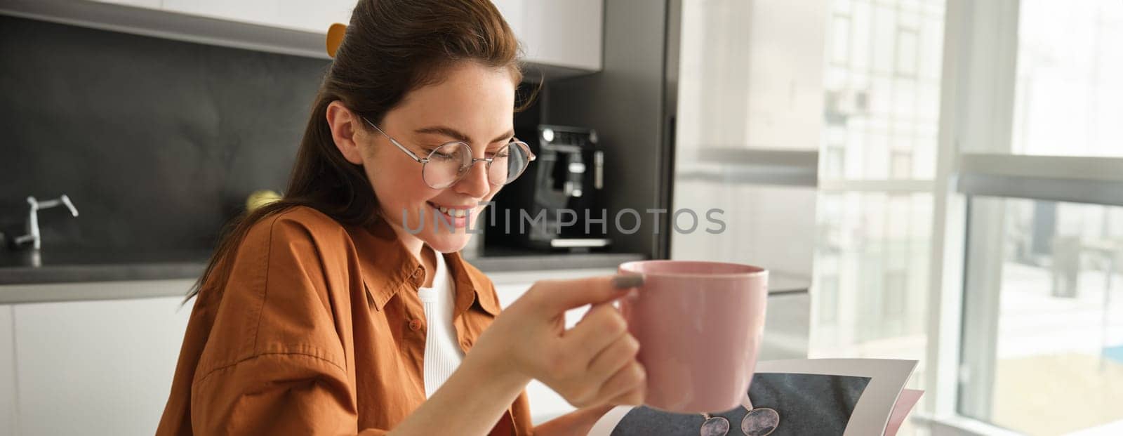 Portrait of young beautiful woman, relaxing with cup of fresh aroma coffee, holding mug and sitting in kitchen, smiling from happiness.