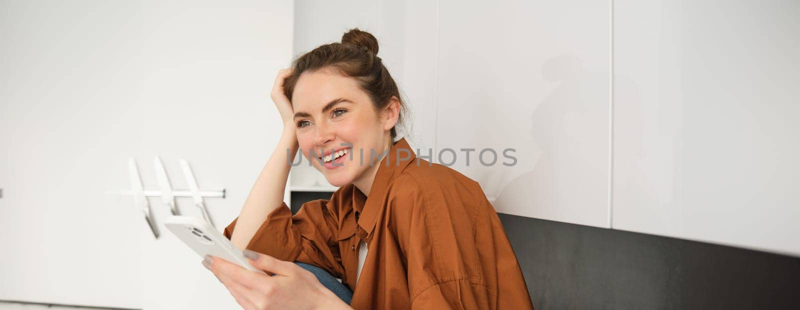 Portrait of young woman with mobile phone, browsing social media, online shopping, sitting on kitchen counter, smiling and looking happy by Benzoix