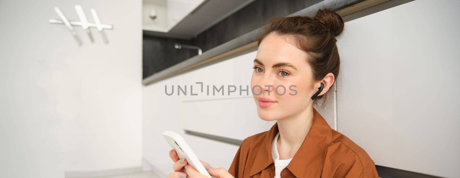 Carefree smiling woman with smartphone, messaging, using mobile phone, sitting on kitchen floor, listening music in wireless headphones.