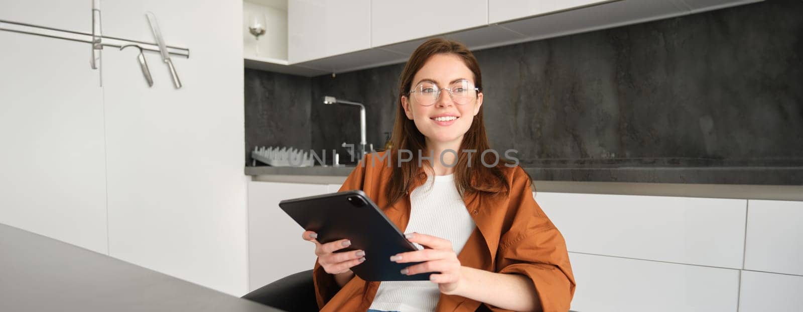 E-learning and remote workplace concept. Young woman in glasses, student studying at home in kitchen, reading on digital tablet, looking at her project, connects to online meeting.