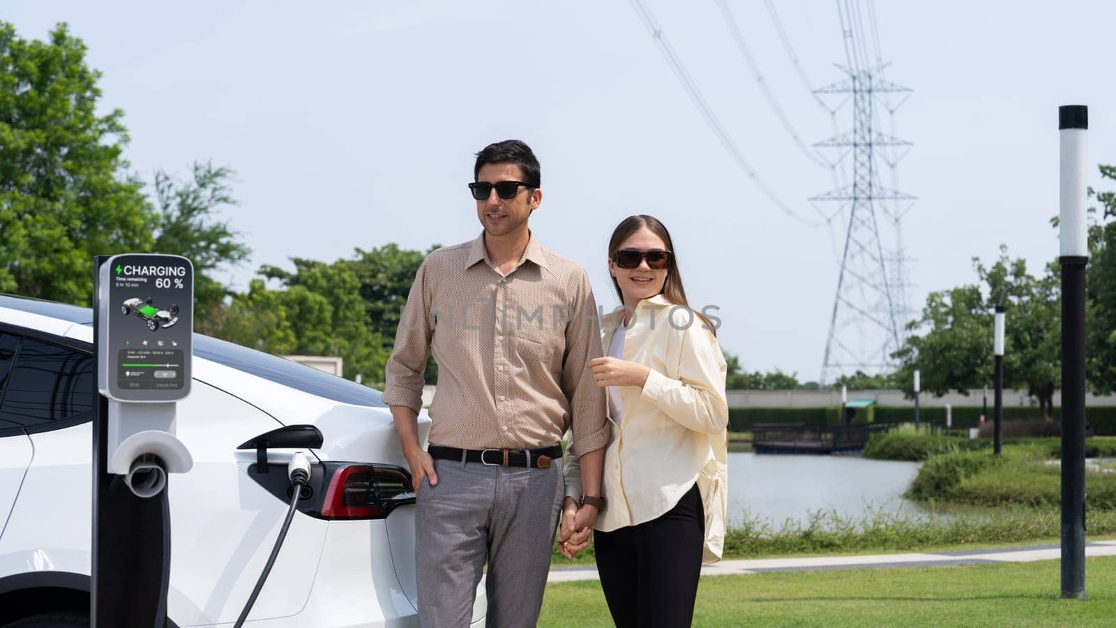 Young couple recharge EV car battery at charging station connected to power grid tower electrical industrial facility as electrical industry for eco friendly vehicle utilization. Expedient