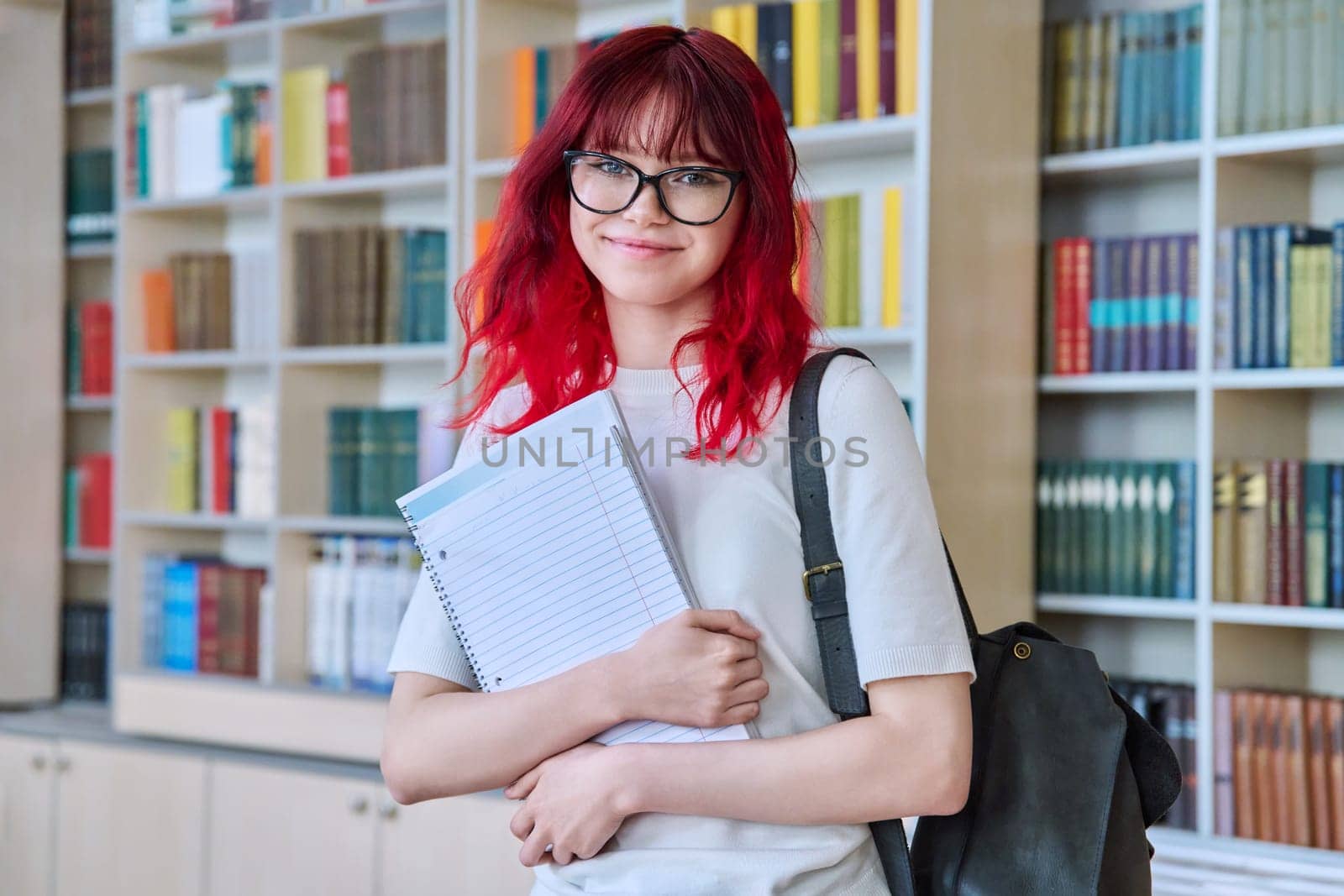 Portrait of teenage female student looking at camera in library. Smiling beautiful girl teenager wearing glasses, holding notebook backpack. College, university, education, knowledge, youth concept
