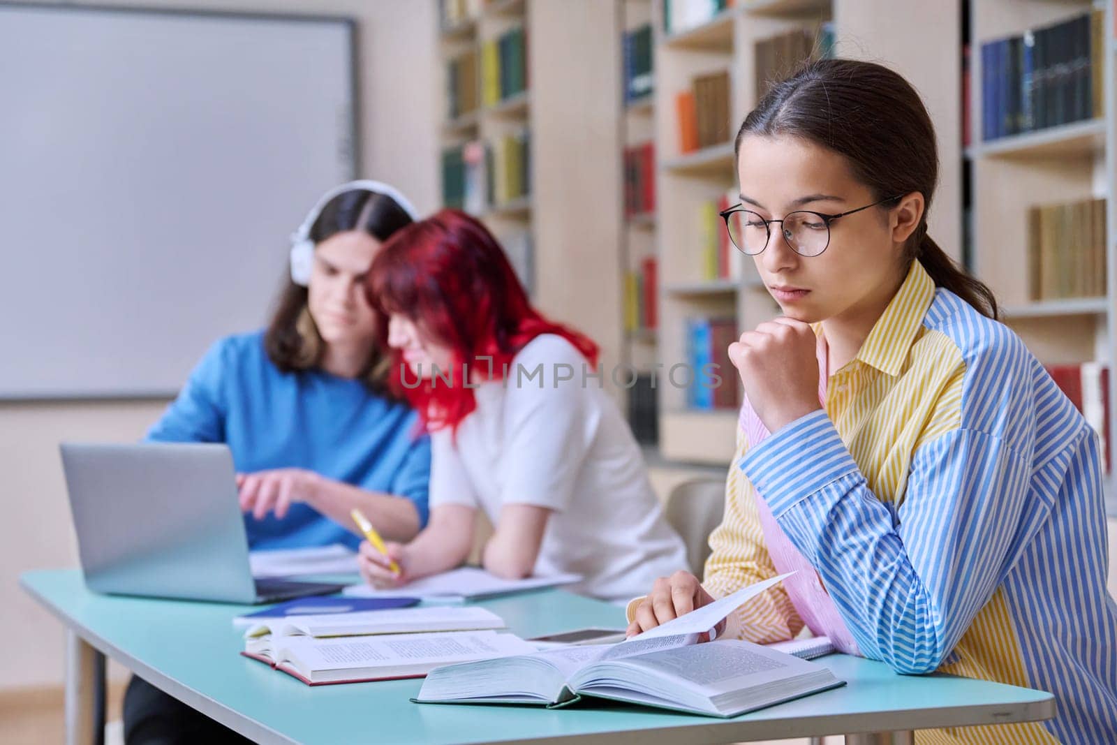 High school students studying in library class, teenage girl in focus. Group of teenagers sitting at desk, writing in notebooks, using laptop books. Knowledge, education, adolescence concept