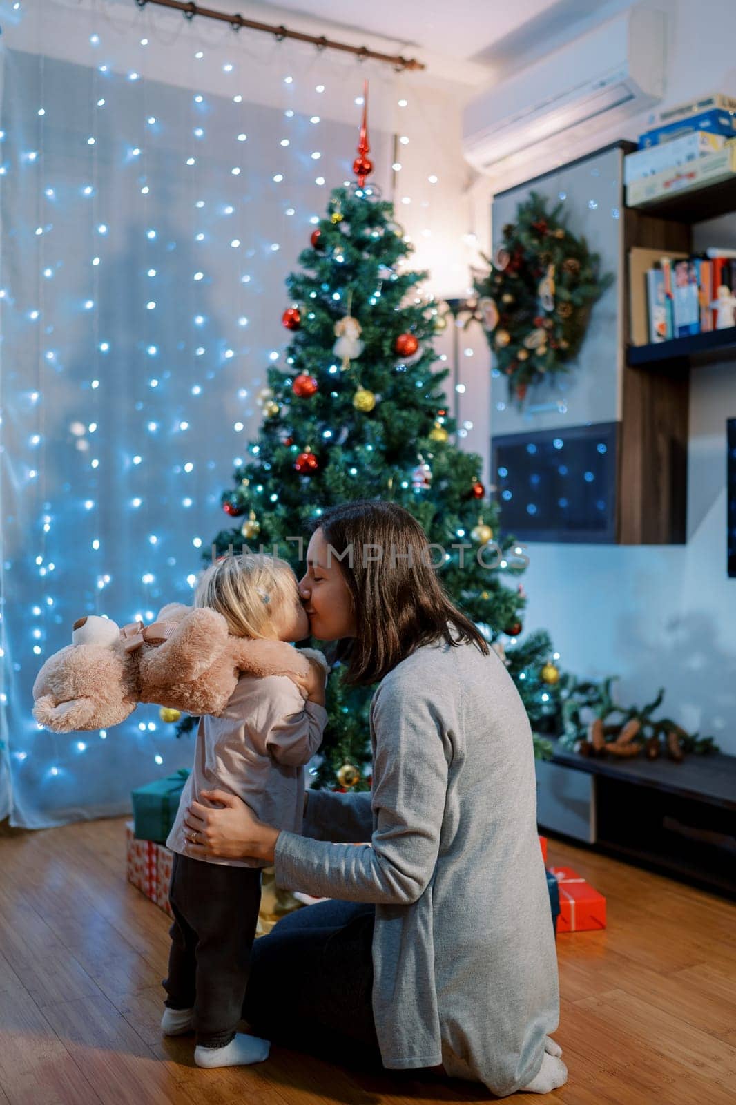 Mom kisses on the cheek a little girl with a teddy bear on her shoulders sitting near a sparkling Christmas tree. High quality photo