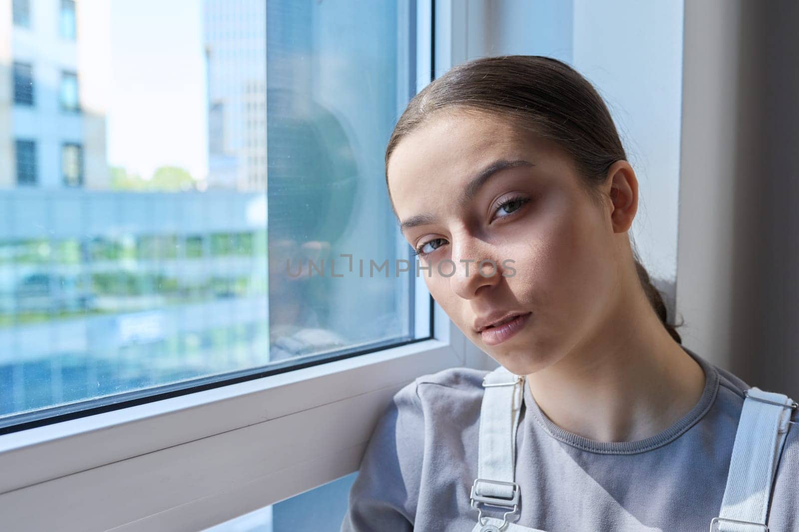 Headshot portrait of young teenage serious girl looking at camera, at home near window. Adolescence, high school, lifestyle, youth, beauty, teenagers concept
