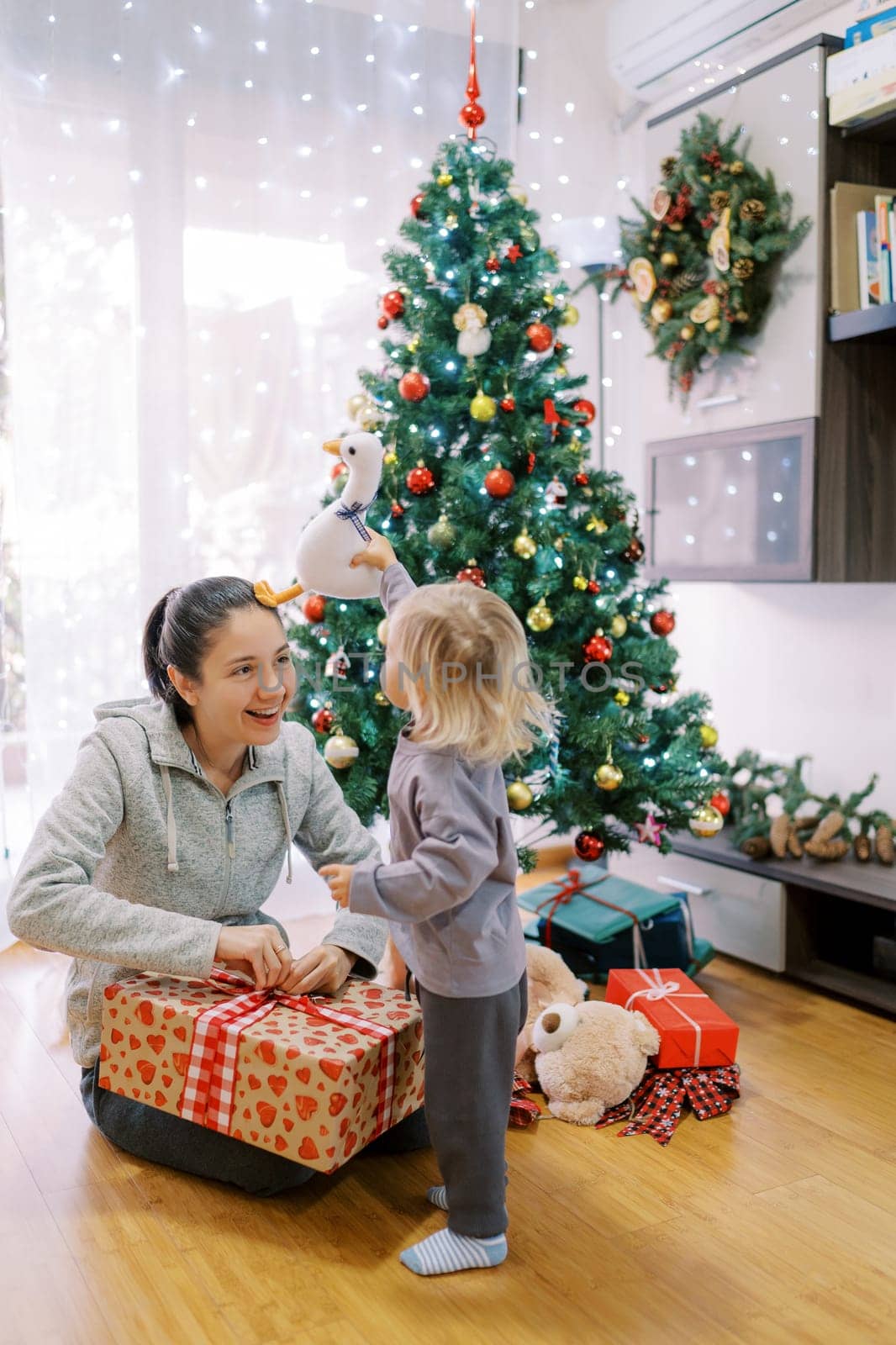 Little girl touches her mother head with a toy duck while she unpacks a gift near the Christmas tree by Nadtochiy