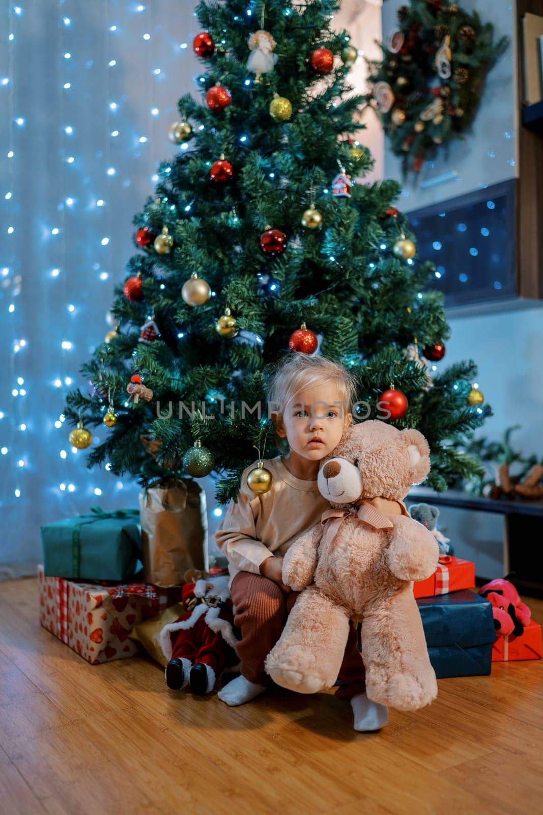 Little girl sits on gifts under the Christmas tree, hugging a teddy bear by Nadtochiy