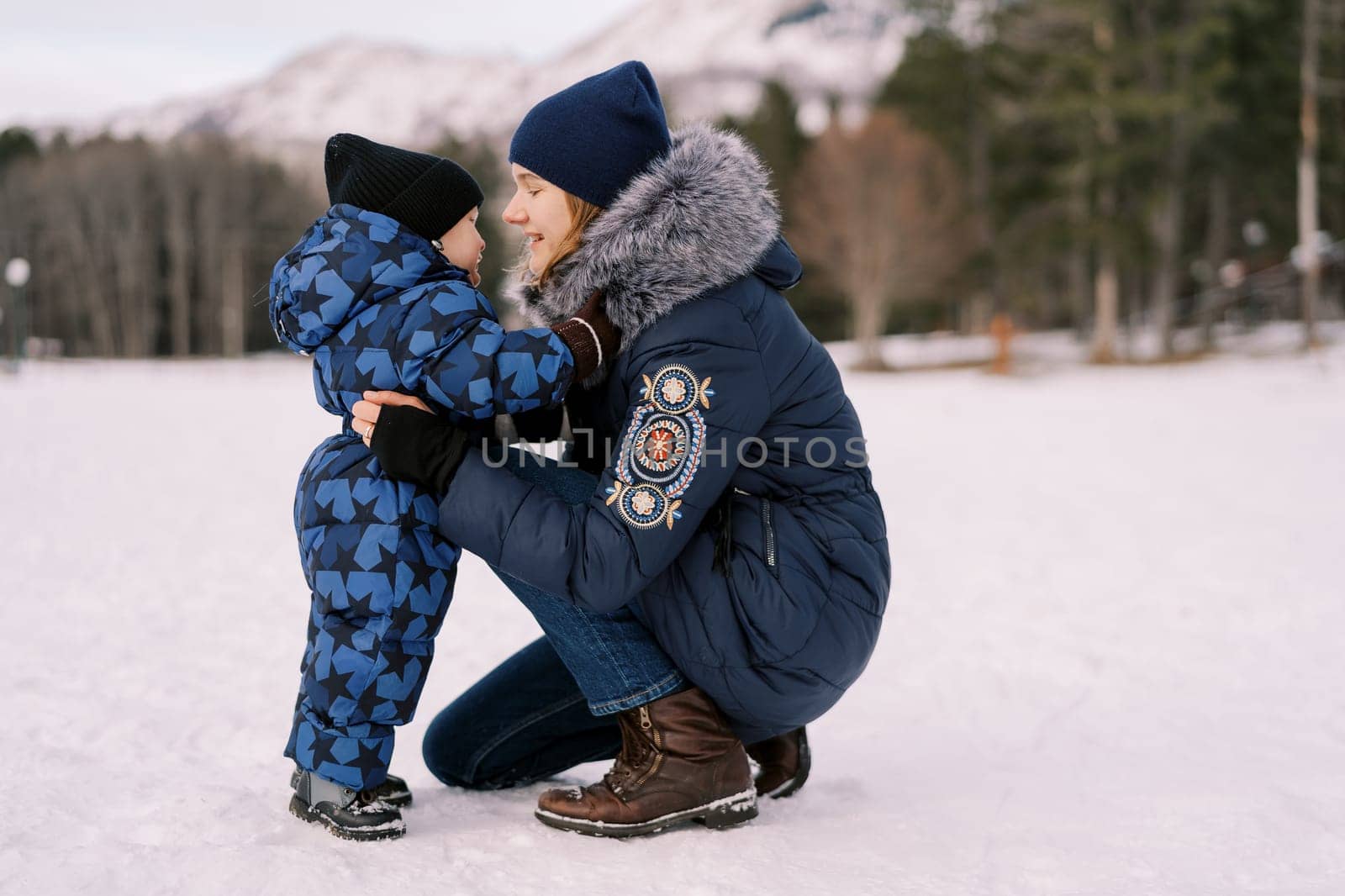 Smiling mother squatted down hugging a little boy in a snowy clearing. Side view by Nadtochiy