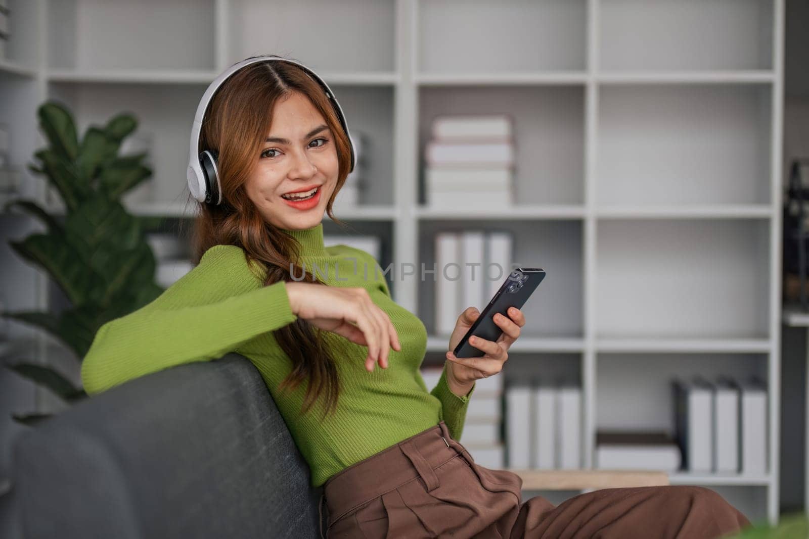 Happy Asian woman listening to music on mobile phone while sitting on sofa at home Relaxed smiling girl with headphones in the morning It's time to rest..