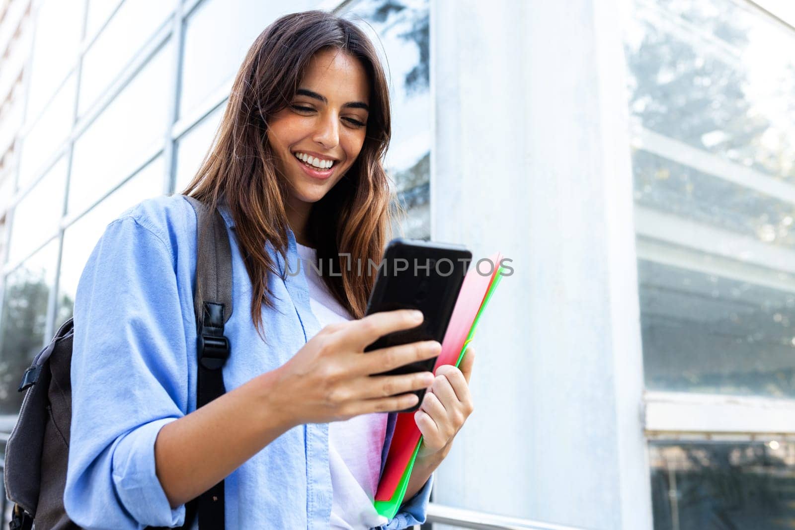 Happy young female university student looking mobile phone standing outside university building. Copy space. by Hoverstock