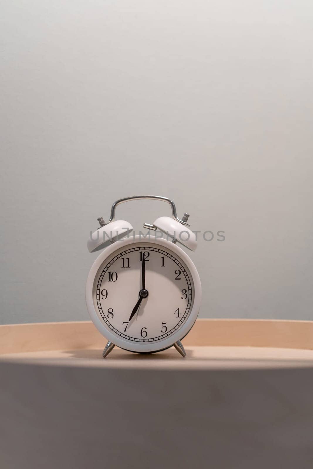 Close-up of a round white alarm clock on a table in the bedroom. The hands on the clock show seven o'clock in the morning, time to get up. Retro alarm clock on the table, vintage tone. space for text