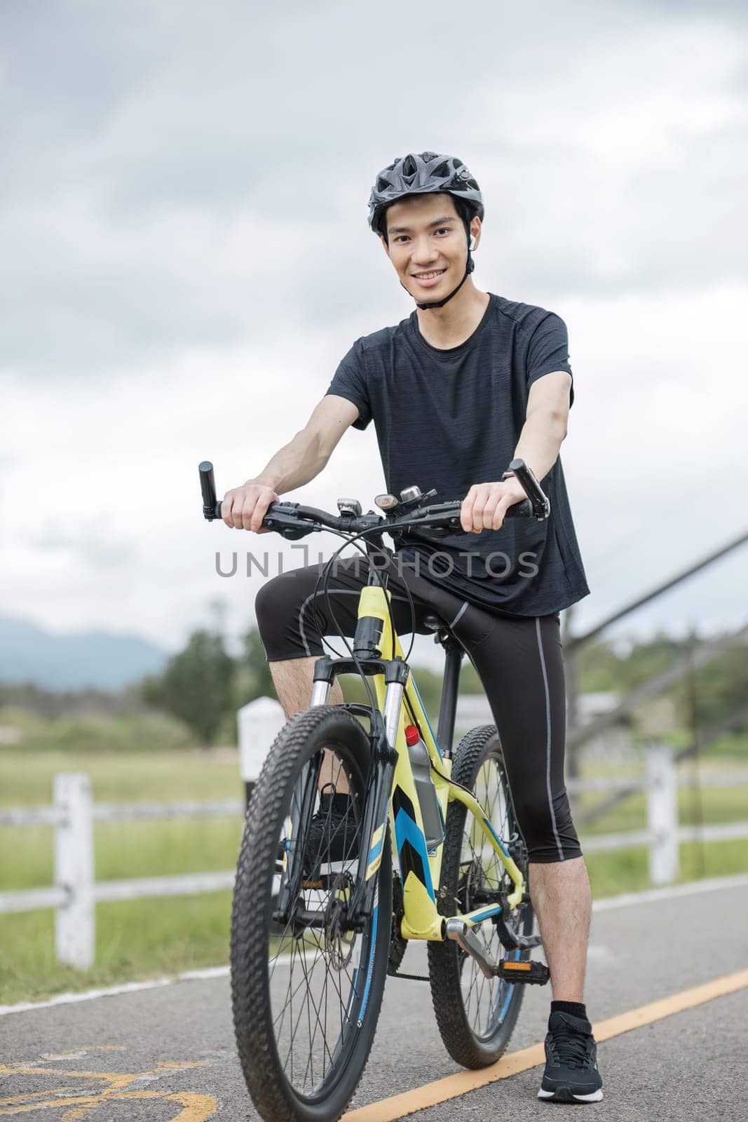 A happy and handsome Asian man in sportswear and a bike helmet, enjoys listening to music through his earbuds while riding a bike on the country roads on the weekend. by wichayada