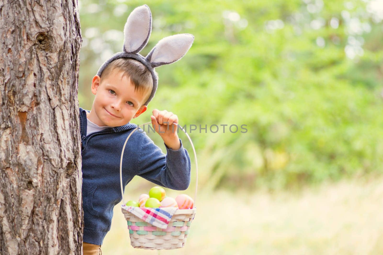 Happy child looks out of a tree holding basket full of colorful easter eggs after egg hunt at spring time. Happy Easter day.