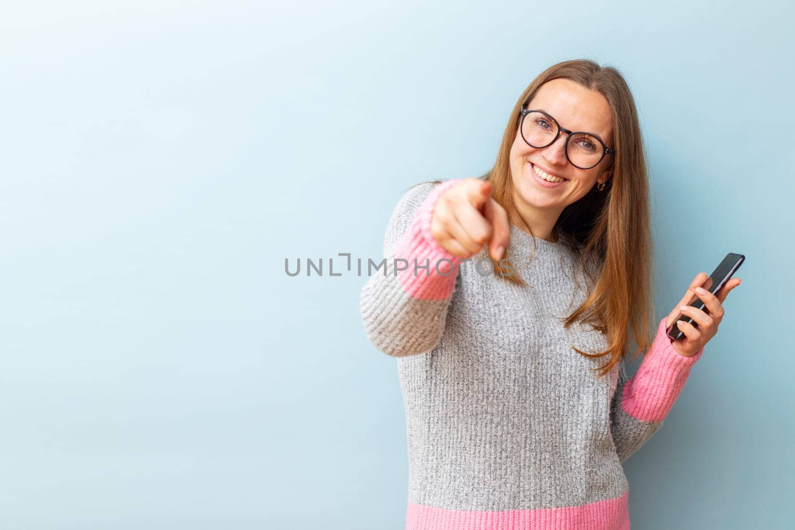 Portrait of happy brunette woman in glasses holding smartphone and pointing her index finger at the camera on blue background.