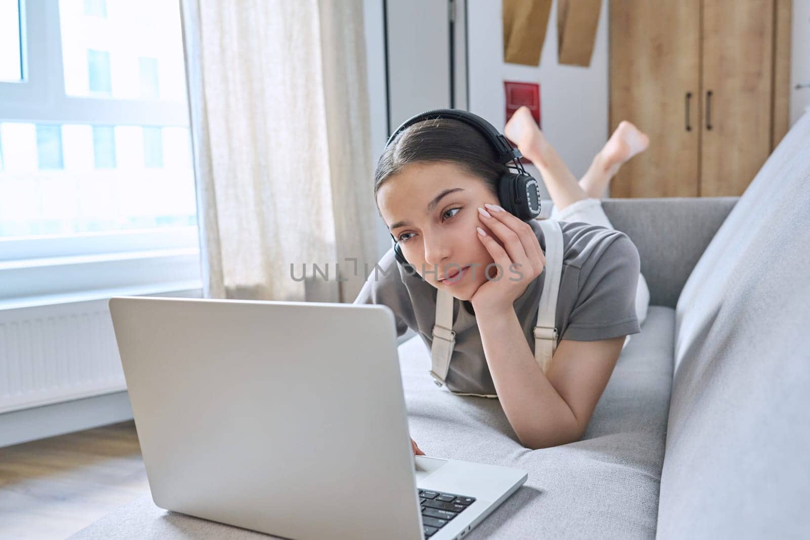Teen girl in headphones at home on couch using laptop by VH-studio