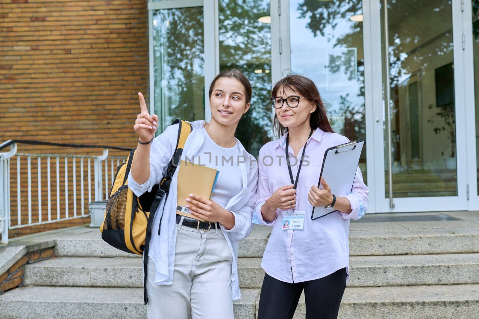 Teenage girl student talking to female teacher, standing on steps of educational building by VH-studio