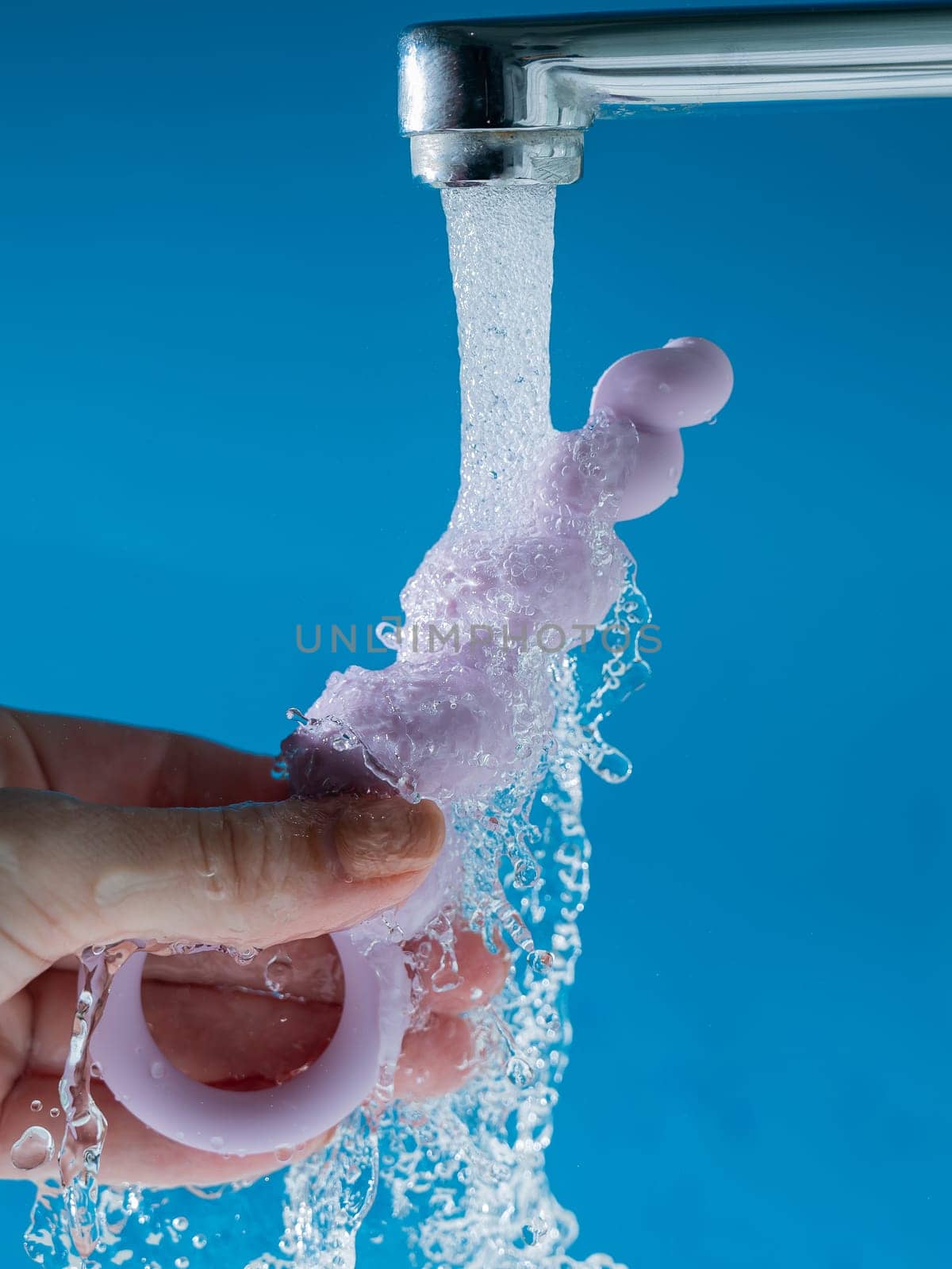 Woman holding lilac anal beads under running water on blue background. Sex toy hygiene concept