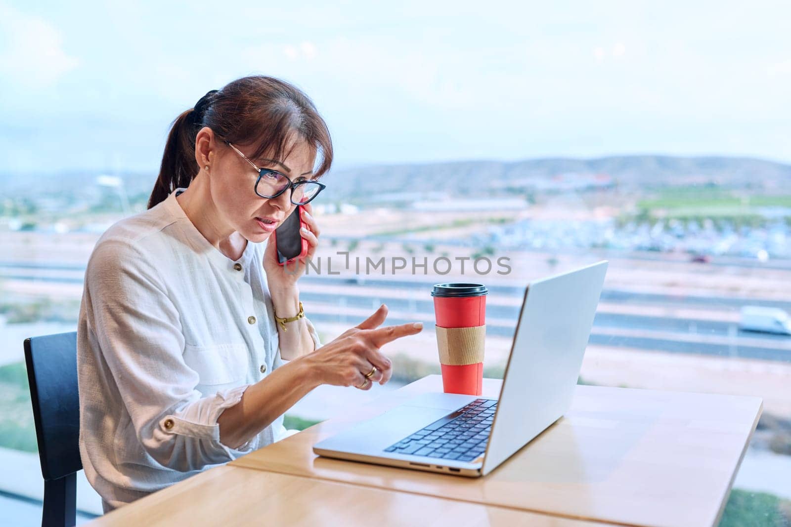 Middle-aged woman in an airport cafe working with a laptop computer, sitting at table with takeaway coffee, near panoramic window