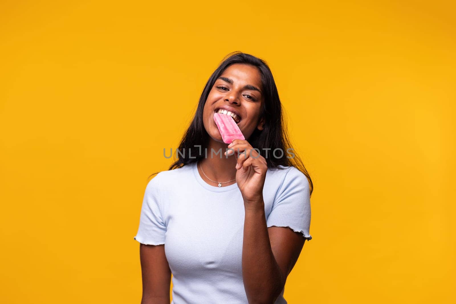 Young Indian woman eating pink popsicle on yellow background. Asian woman eating ice cream. Looking at camera. Studio shot.
