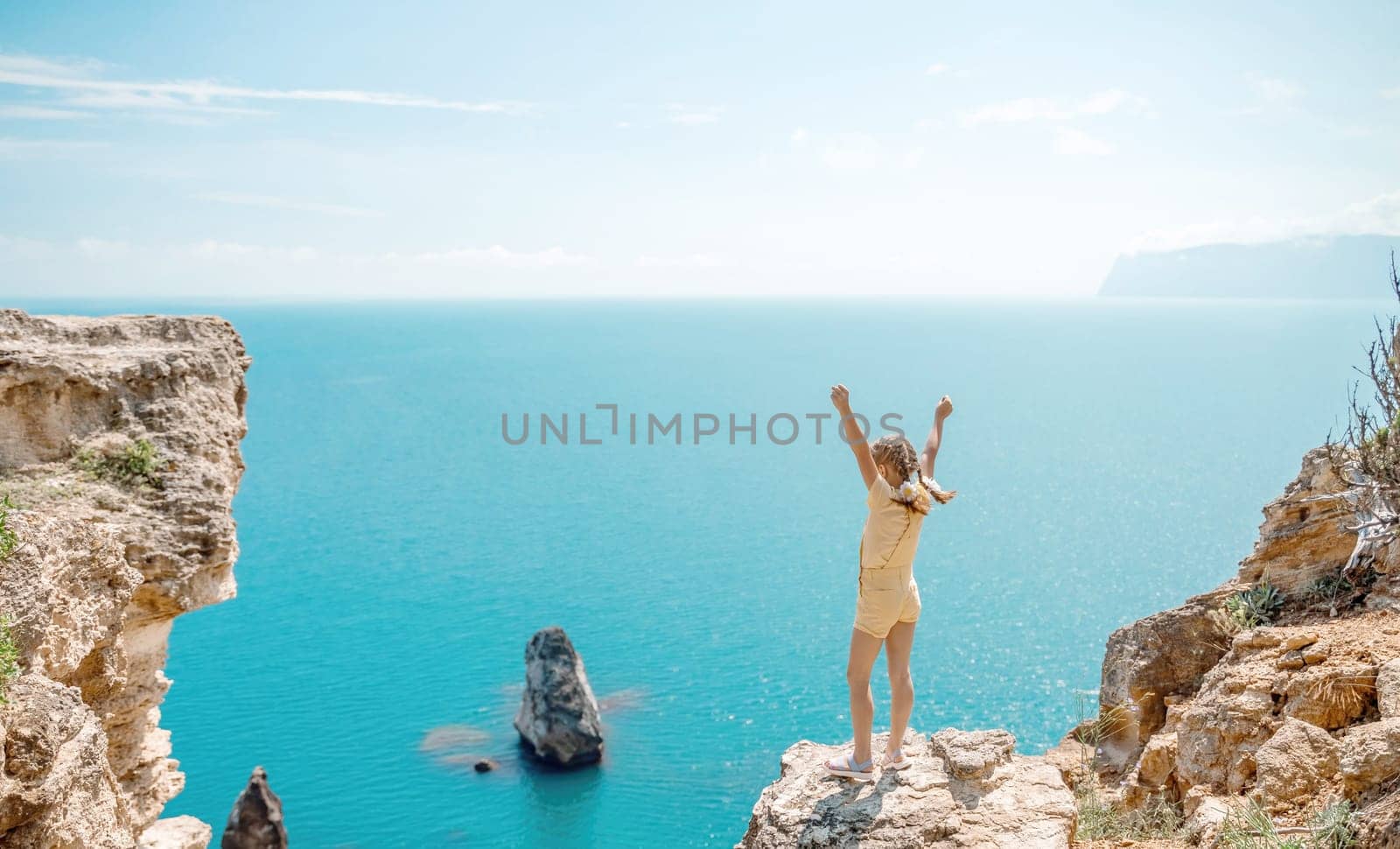 Happy girl stands on a rock high above the sea, wearing a yellow jumpsuit and sporting braided hair, depicting the idea of a summer vacation by the sea. by Matiunina